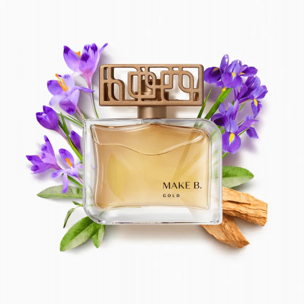 Frasco do Make B Gold transparente tampa ornada em dourado. Atrás do frasco estão flores Violetas. 